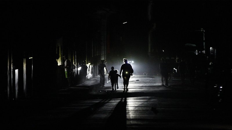 Color photo of a Cuban street in the dark, showing a the silhouettes of people lit up by headlights.