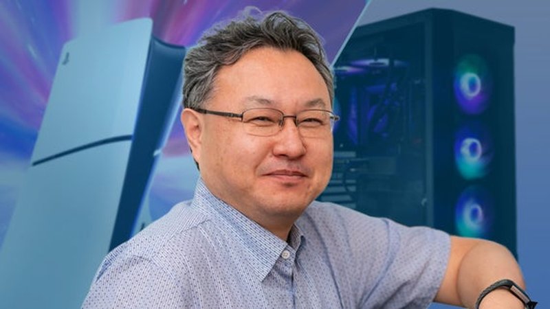 Shuhei Yoshida poses with one arm up in a relaxed manner, behind him a PlayStation 5 console and a PC