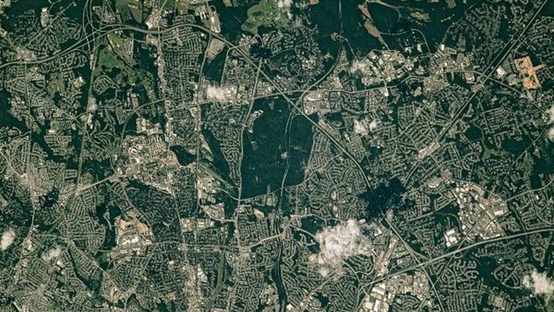 A straight-down view of Greenbelt is centered on a square park, with smaller green spaces weaving through surrounding homes, businesses, a college campus, and government buildings.