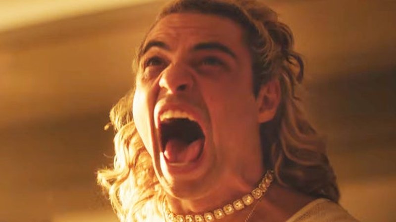 Noah Centineo as Ken in the Street Fighter film. He is screaming and has his blonde hair pulled back in a half bun. He has a choker around his neck