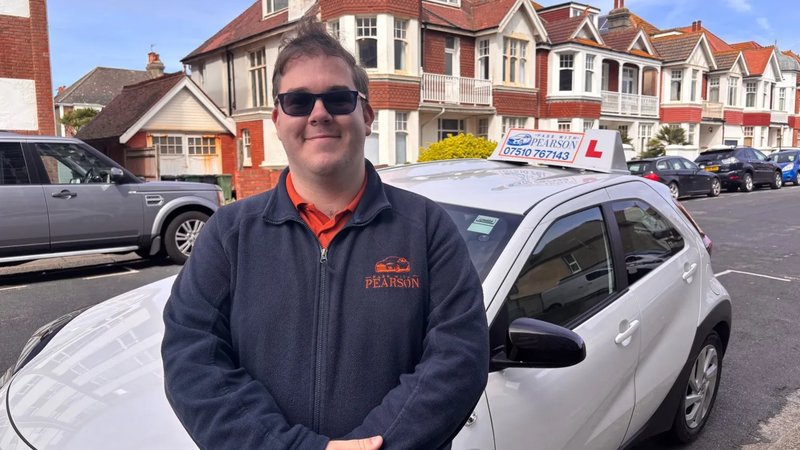 Driving instructor Joe Pearson, wearing sunglasses, smiles as he stands in front of a car.