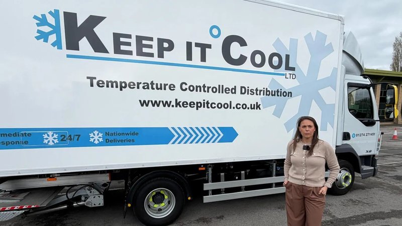A woman in a grey cardigan and brown trousers stands in front of a blue and white van parked on a forecourt.